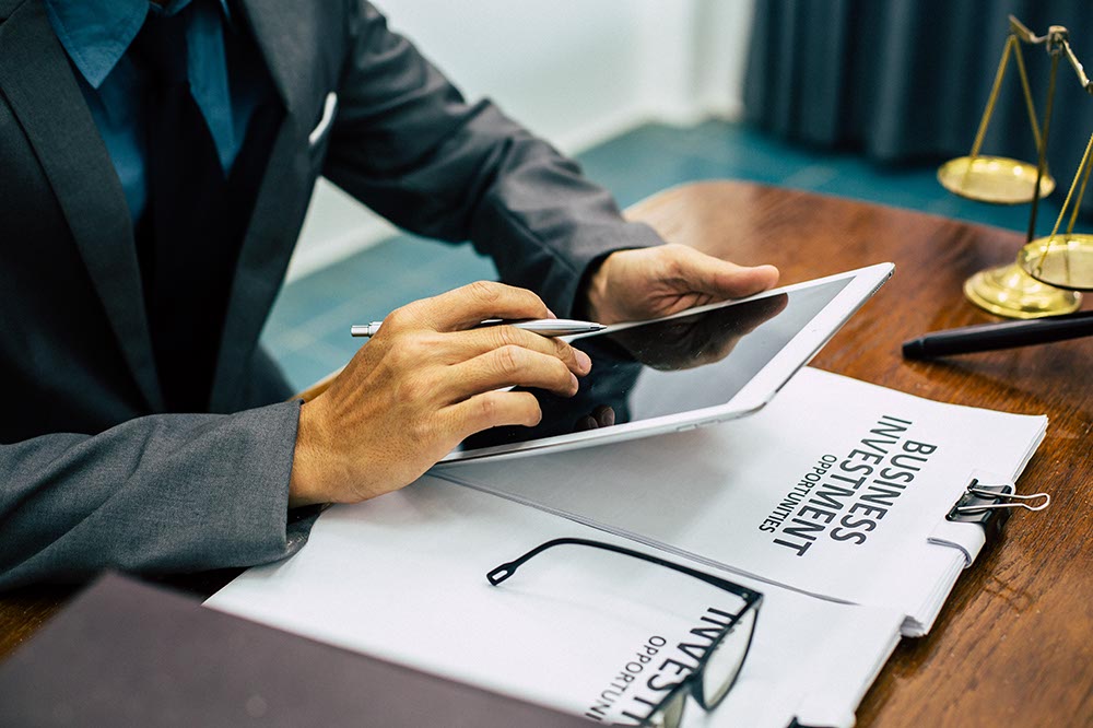 Image of hands holding an iPad with stylus at desk with arms resting on bundle of papers with cover page reading: Business Investment Opportunities