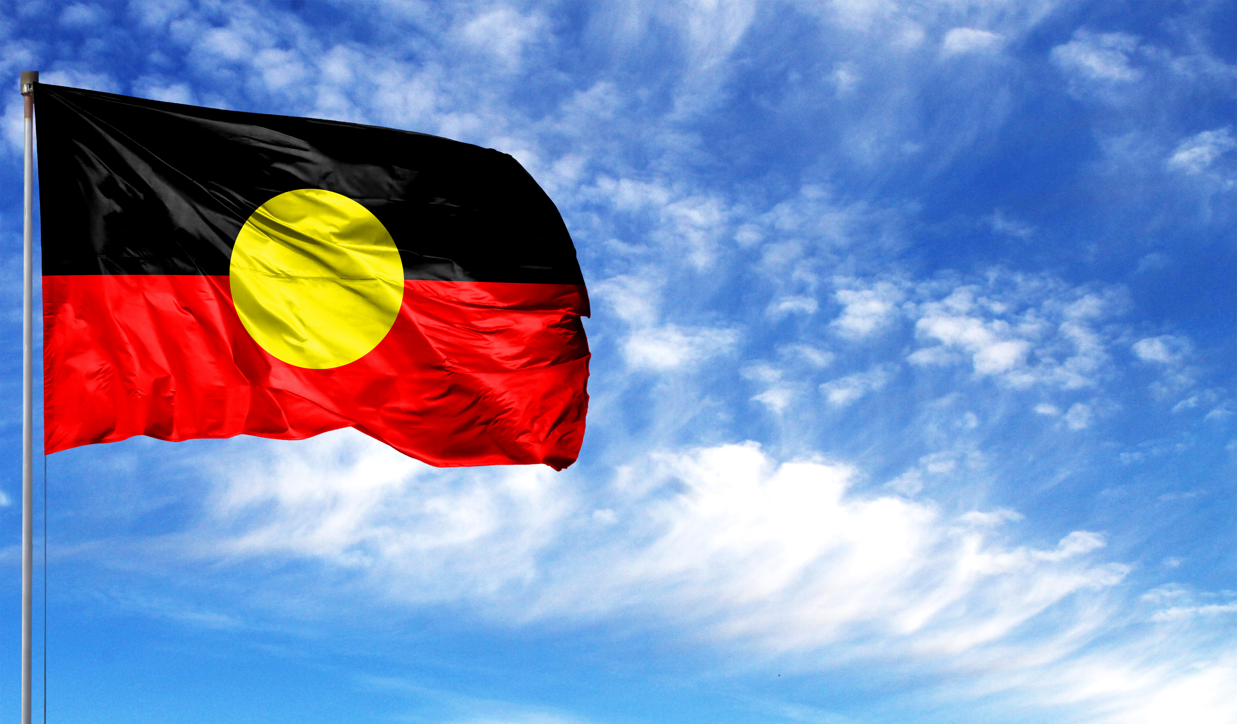 Image of the Indigenous Flag on a flag pole against a blue sky with some white clouds